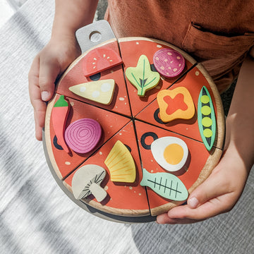  Wooden pizza toy. The set includes 6 slices and a pizza tray with 12 interchangeable toppings held in place by small velcro spots, all enclosed in an illustrated cardboard box. 
